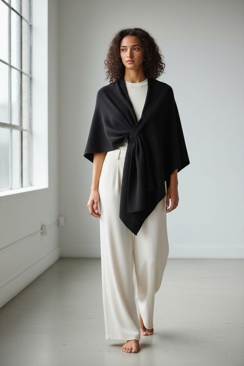 A person with curly hair stands barefoot in a bright, minimal room with large windows, dressed in a white outfit and draped in the MERSEA - MODERN TRAVEL WRAP IN BLACK. This elegant wrap from MERSEA features a securing loop, blending style and functionality for an effortlessly chic look.