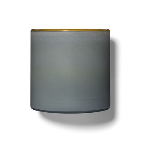 A minimalist cylindrical lamp shade from LAFCO, named SEA DUNE BEACH HOUSE 15OZ, features a smooth gray surface with a slight gold trim on the top edge, casting a subtle shadow reminiscent of sea spray on a plain white beach house background.
