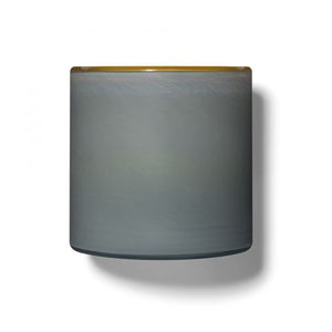 A minimalist cylindrical lamp shade from LAFCO, named SEA DUNE BEACH HOUSE 15OZ, features a smooth gray surface with a slight gold trim on the top edge, casting a subtle shadow reminiscent of sea spray on a plain white beach house background.