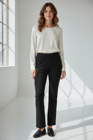 A woman stands indoors by a large window, wearing a long-sleeve cream blouse and the KRAZY LARRY, INC PULL ON WIDE ANKLE PANT in black, which features a tummy tuck design. She completes her look with black loafers and smiles slightly, her wavy brown hair catching the sunlight that casts shadows on the white floor and wall.