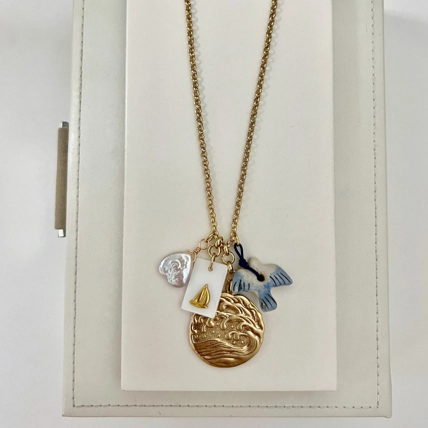 The SANDRINE FRANCE STUDIO WREN NECKLACE is a handmade gold necklace from NYC featuring four charms—a white shell, a rectangular gold sailboat tag, a gold wave pendant, and a blue-white fish—on a white jewelry stand.
