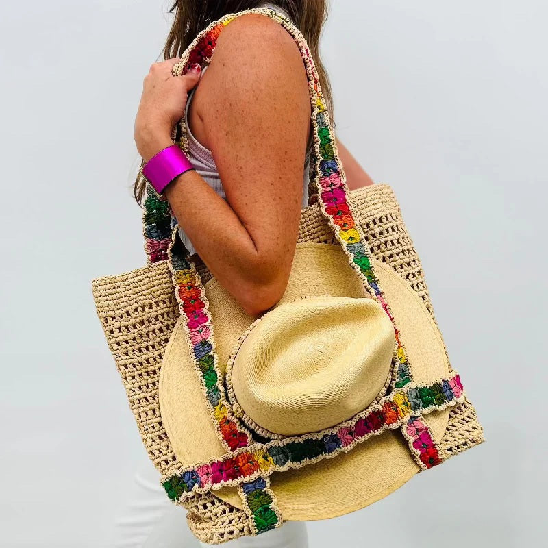 A woman holds the BALDIZ INDIE Raffia Tote with rainbow straps, paired with a straw hat. She wears a purple bracelet and a sleeveless white outfit, standing against a plain background.