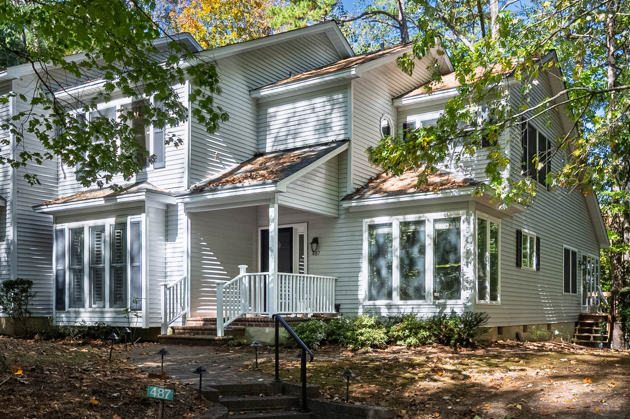 A two-story white house with large windows, a small front porch with white railings, surrounded by trees and greenery, with dappled sunlight filtering through the leaves.