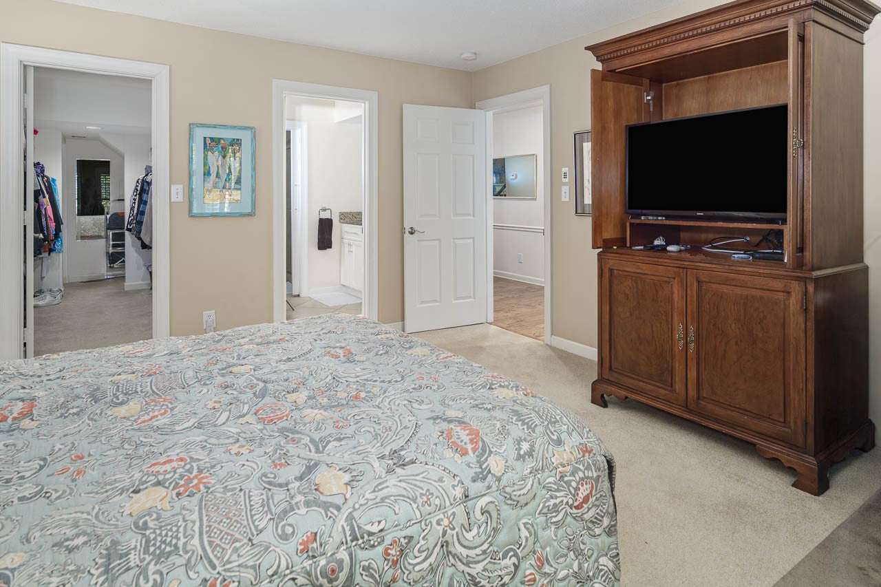 A bedroom with a bed featuring a patterned comforter, a wooden TV cabinet with a flat-screen TV, beige carpet, doors leading to a bathroom, hallway, and walk-in closet, and artwork on the wall.