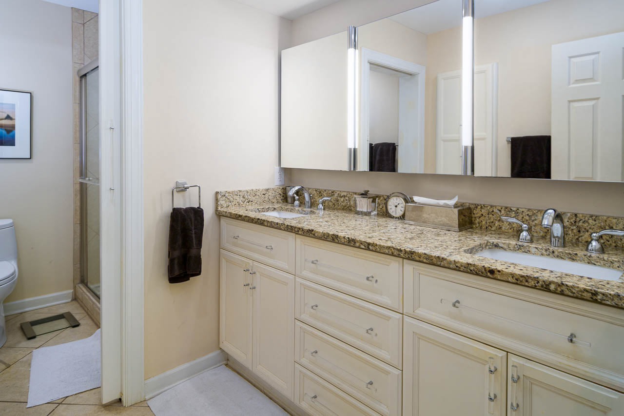 A modern bathroom with a double-sink granite countertop, white cabinets, large mirrors with vertical lights, a towel hanging on a rack, and a toilet and glass-enclosed shower visible in the background.
