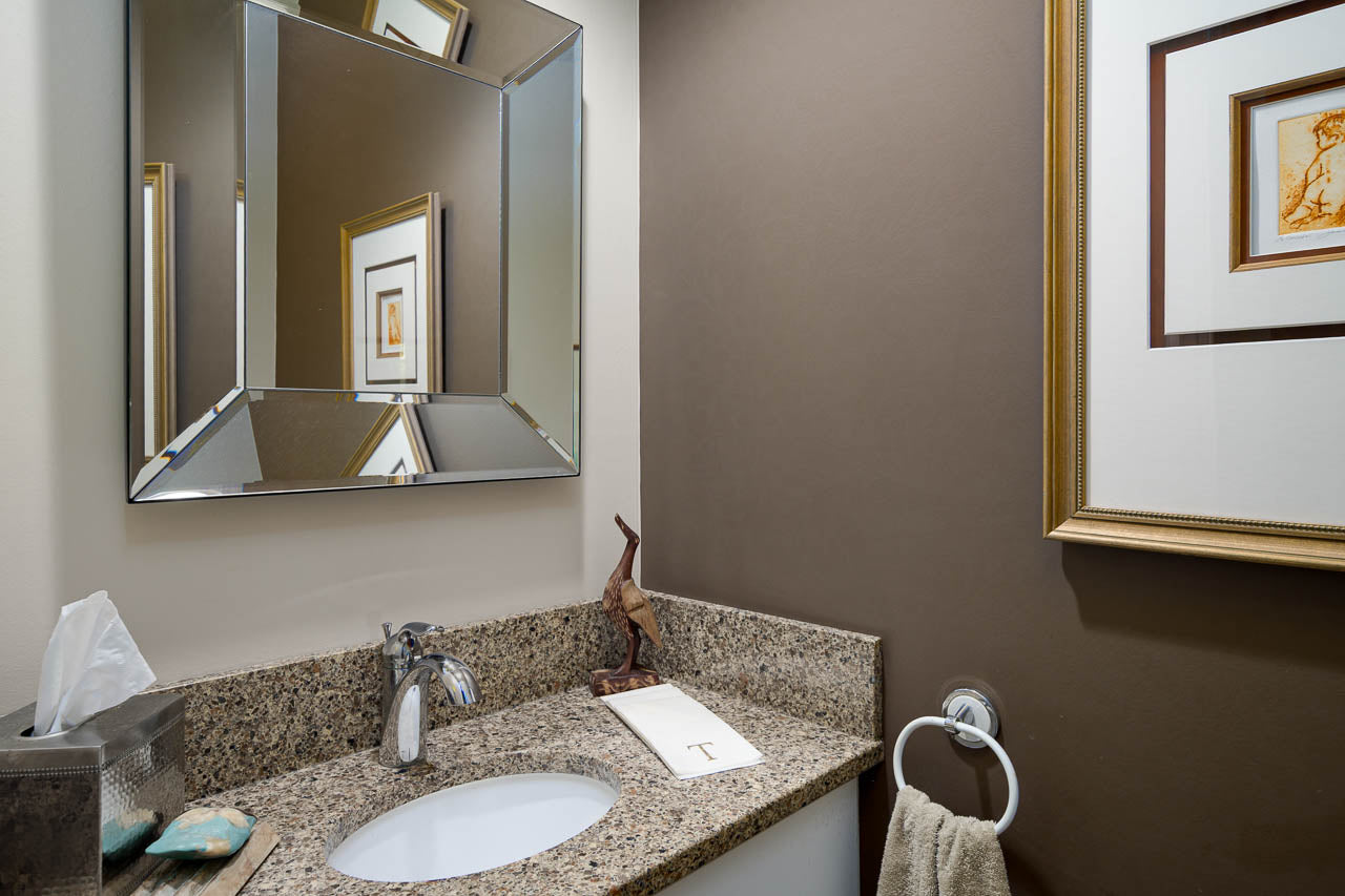 Small bathroom with a granite countertop, modern faucet, and geometric mirror above the sink. Towel hangs on a ring, framed art on the wall, and a tissue box and decor piece sit on the counter.
