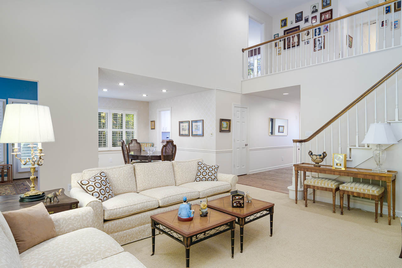 Bright living room with cream-colored sofas, patterned cushions, wooden coffee table, and decorative lamps. Open layout leads to dining area. Staircase and gallery wall with framed photos are visible. Neutral tones throughout.