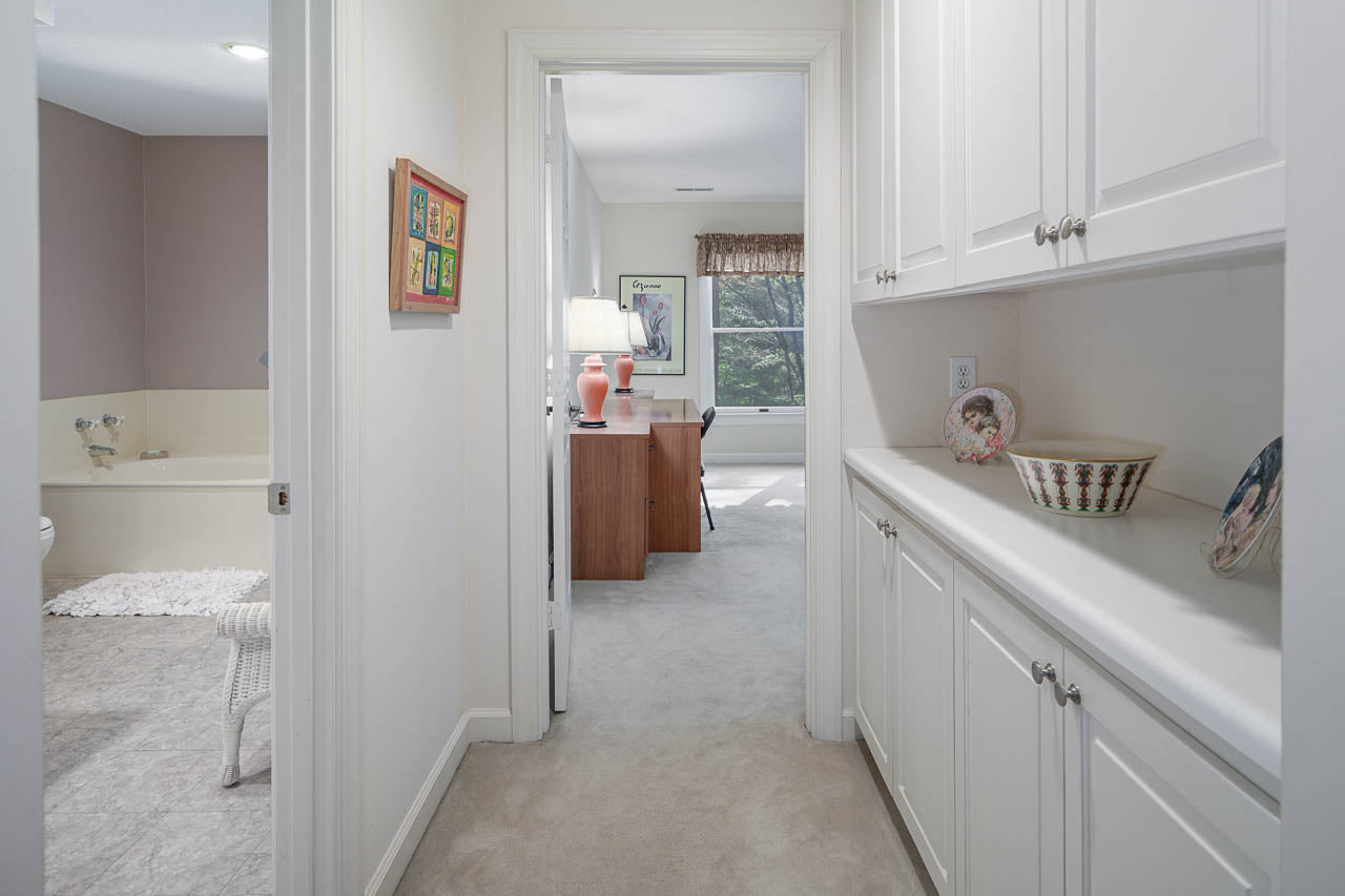 Hallway with white cabinets on the right, leading to a bright bedroom with a desk and lamp by the window; a bathroom with a bathtub is visible to the left. Decorative items sit on the cabinets.