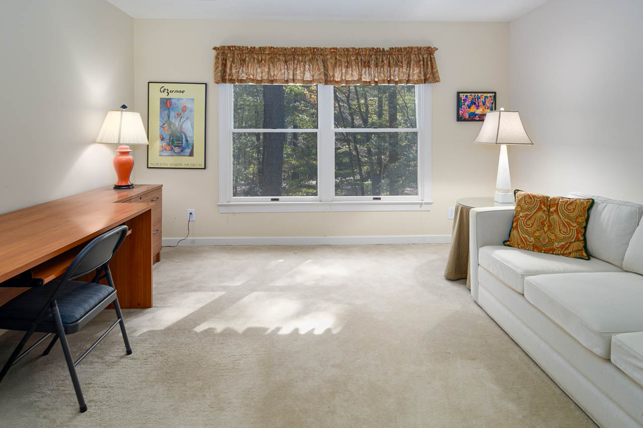 A bright room with beige carpet, a white sofa with patterned pillows, a wooden desk with a chair and lamp, two lamps, wall art, and large windows showing trees outside.