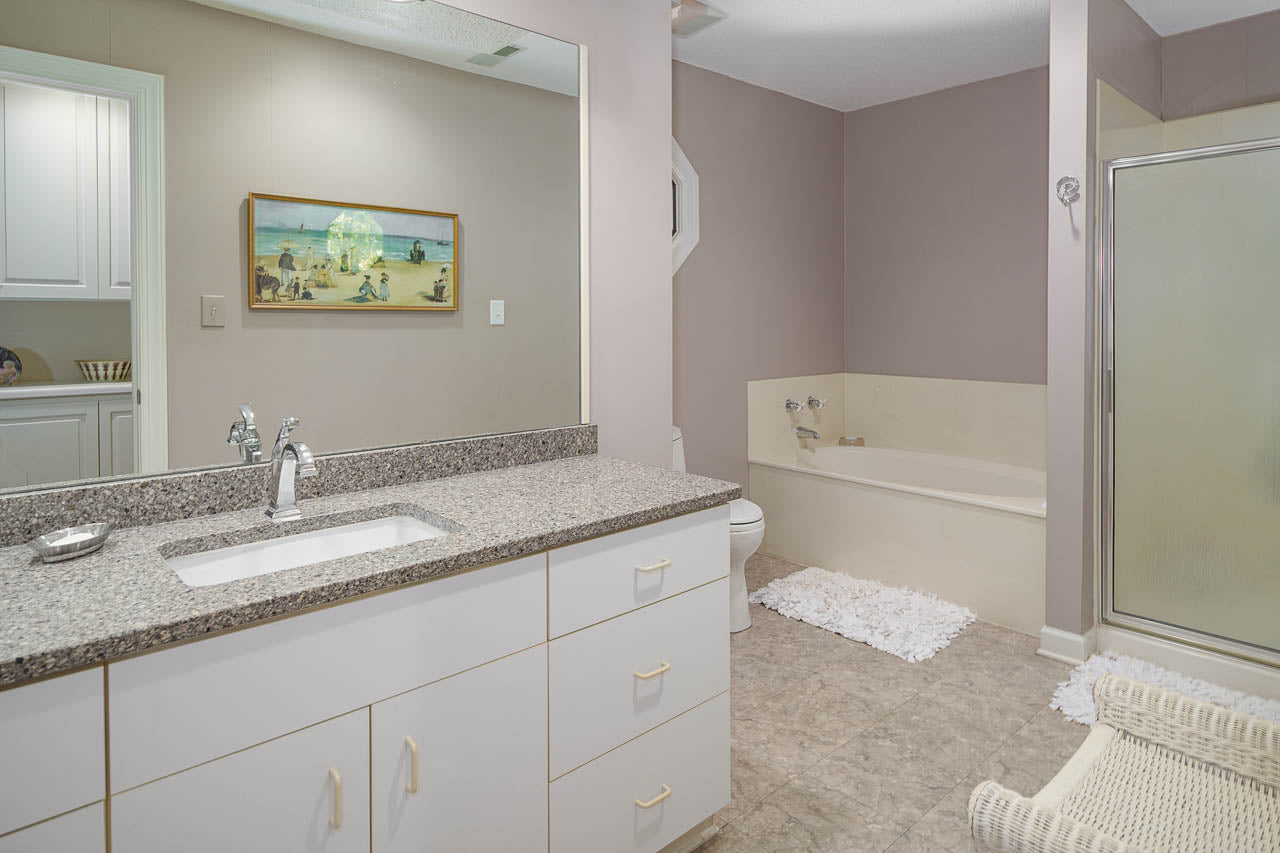 Modern bathroom with granite countertop, white cabinets, a large mirror, bathtub, glass shower, and beige walls. A framed painting hangs above the toilet. Textured floor tiles and white rugs are also visible.