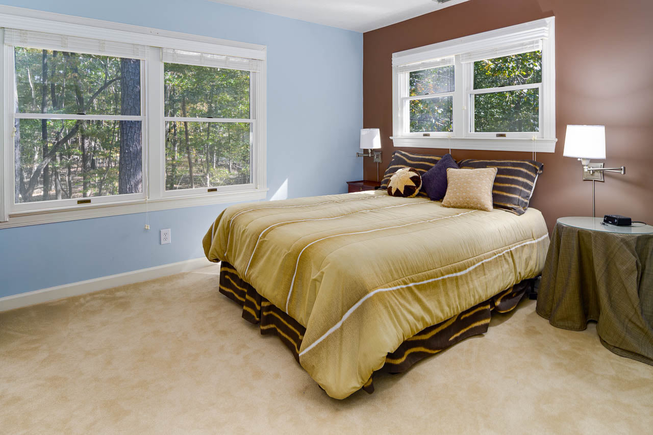 A cozy bedroom with beige carpet, a bed with striped gold and brown bedding, pillows, two nightstands with lamps, light blue and brown walls, and large windows letting in natural light with trees visible outside.