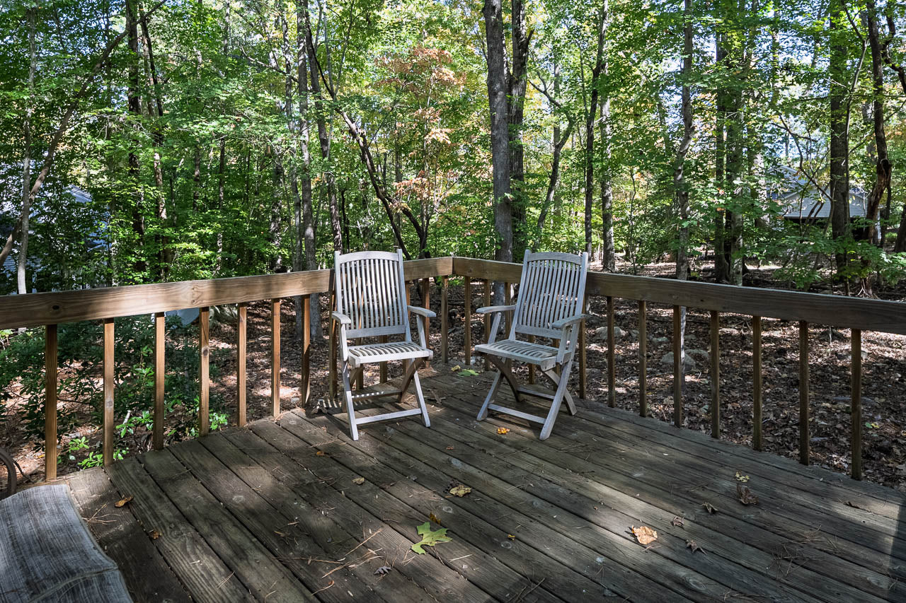 Two wooden chairs sit on a weathered wooden deck surrounded by a forest of tall trees with green leaves and scattered sunlight. A wooden railing encloses the deck, and fallen leaves lie on the deck floor.