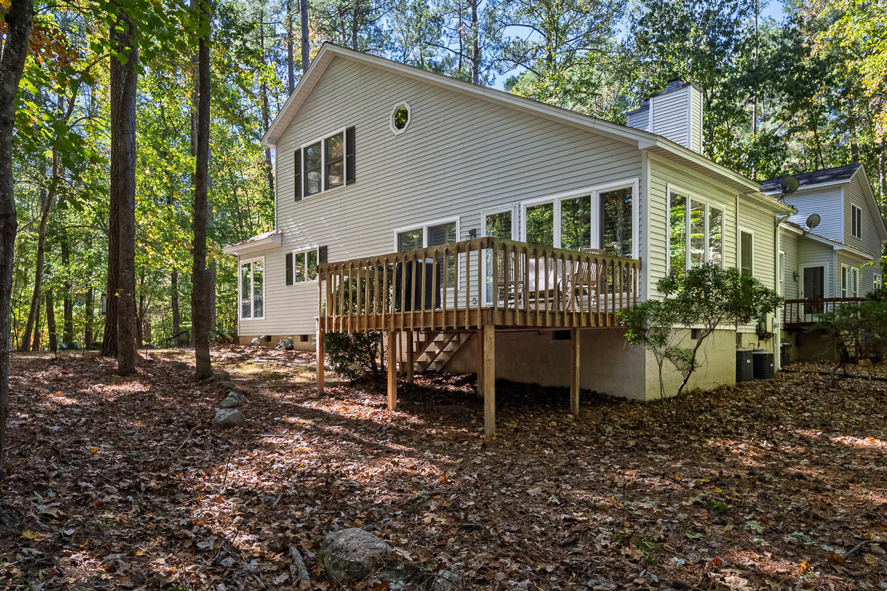 A white house with large windows and a wooden deck stands among tall trees in a sunlit, leafy forest yard with a ground covered in dry leaves.