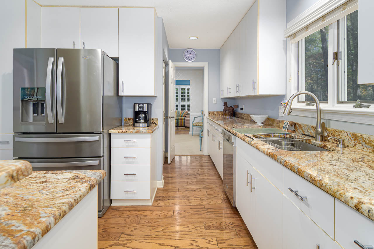 Modern kitchen with stainless steel refrigerator, white cabinets, granite countertops, a coffee maker, sink by a window, and hardwood floors. A doorway leads to another room with a chair and blue accents.