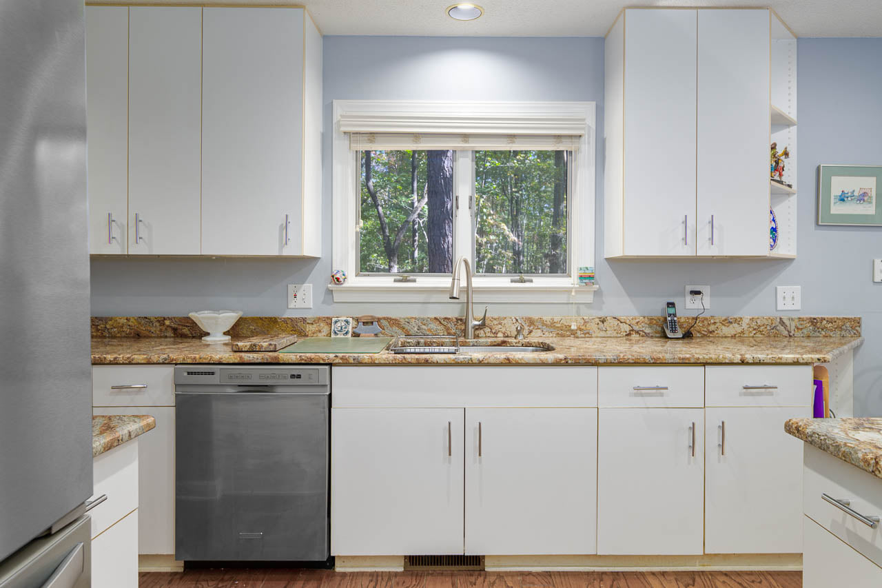 A modern kitchen with white cabinets, granite countertops, a stainless steel dishwasher, and a large window above the sink with a view of trees outside.