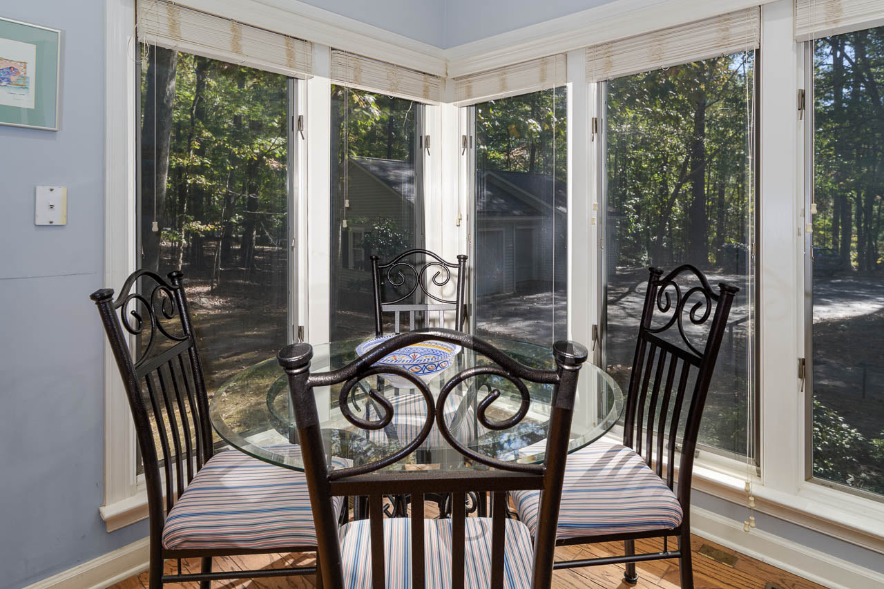 A round glass dining table with four striped-cushion chairs in a sunlit corner, surrounded by large windows overlooking a wooded backyard.