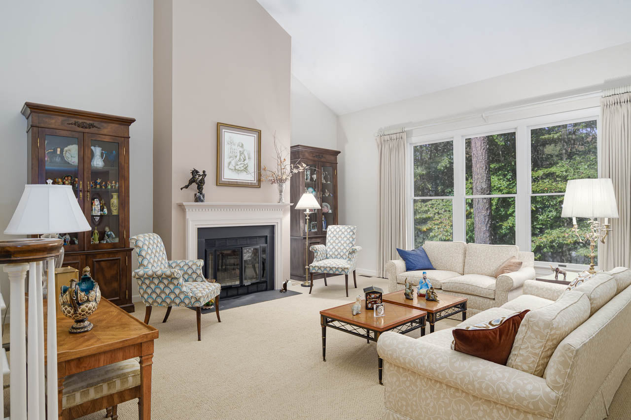 A bright, elegant living room with cream-colored sofas, patterned armchairs, a coffee table, a fireplace, and large windows letting in natural light. Glass display cabinets flank the fireplace, and the room has neutral decor.