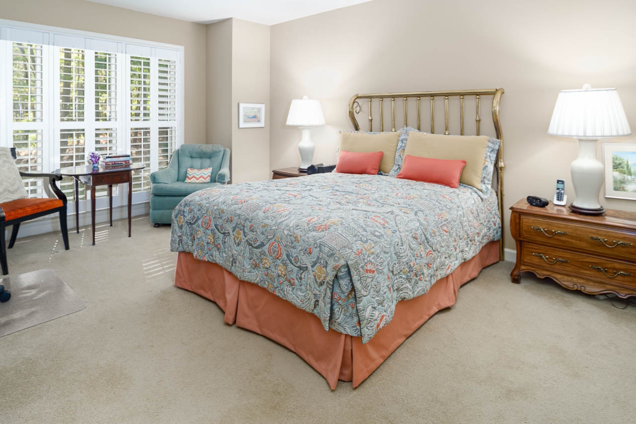 A cozy bedroom with a patterned bedspread, coral bed skirt, brass headboard, two wooden nightstands with lamps, an armchair, a desk with a chair, and large windows with white shutters.