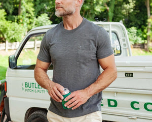 A man with a gray beard wearing the CRIQUET - SLUB COTTON SHORT SLEEVE POCKET T-SHIRT IN WASHED BLACK holds a canned drink in a green koozie outdoors, with trees and a white vehicle blurred in the background.