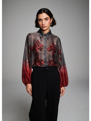 A person with short dark hair wears the MILVA MI - FLORAL RED BLOUSE by MILVA—featuring a gray and red floral pattern—and black high-waisted trousers, standing against a plain gray background.