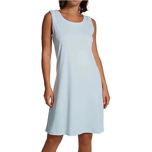 A woman with long hair wearing a simple, sleeveless P. JAMAS - Butterknit Sleeveless Short Gown by FASHIONVIEWS stands against a plain background. The knee-length white dress, made of soft pima cotton knit, features a round neckline and her arms relax by her sides.