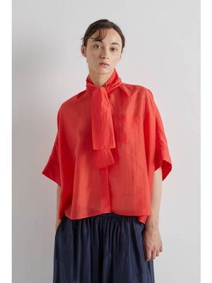 A person in profile wears the MEIMEIJ - SCARF TIE NECK BLOUSE by MEIMEIJ in bright red with short sleeves, paired with a dark, flowing skirt. Their dark hair is tied back, hands in pockets, against a plain white background.