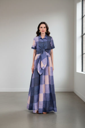A woman wearing the LAVI - FRESIA Tie Front Cropped Top in Quadri, featuring a blue and light purple plaid pattern with short sleeves, a buttoned front, and an oversized tie-front detail reminiscent of a plaid shirt, stands against a plain white background.