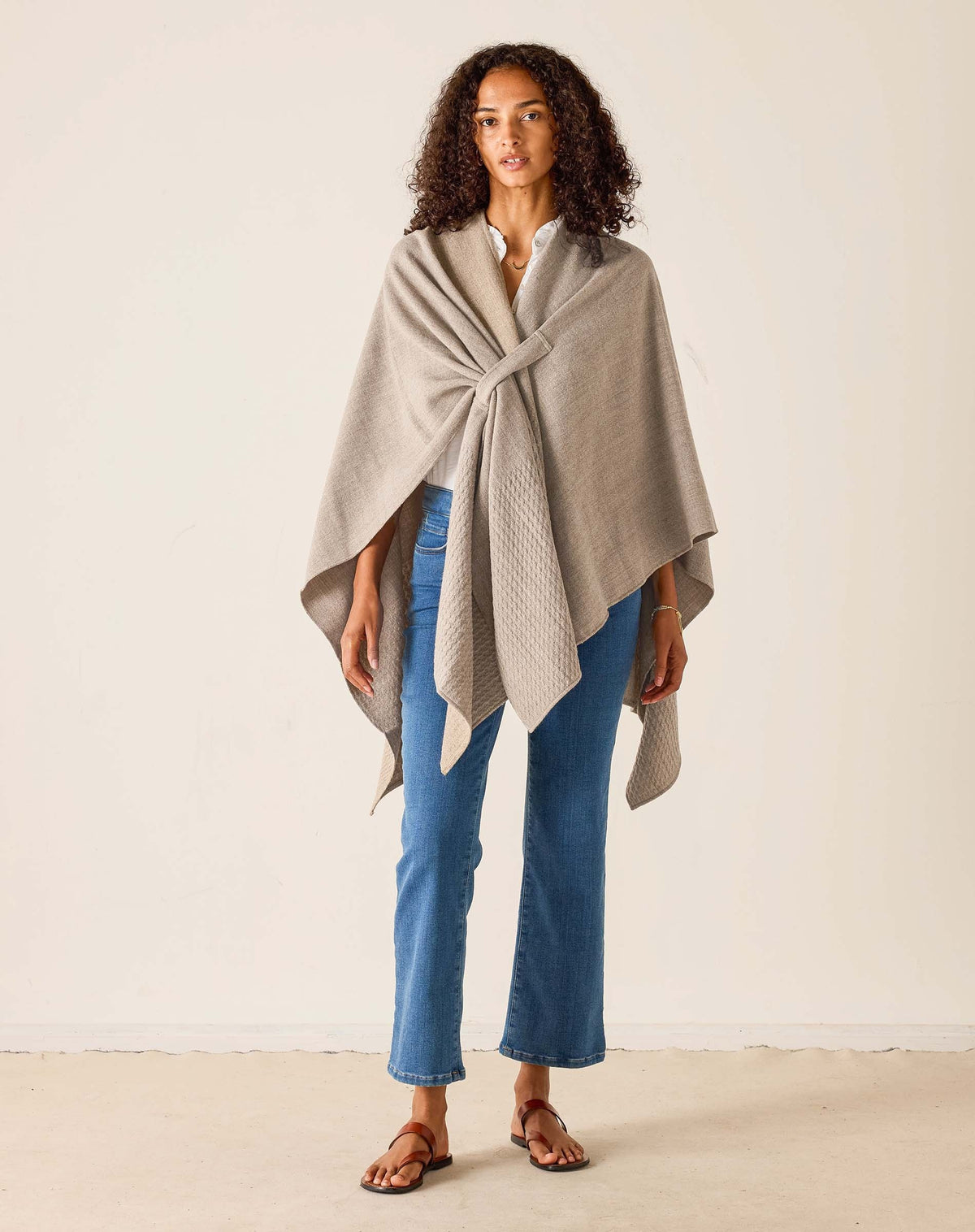 A woman stands indoors in a bright room with light streaming through a large window, smiling softly. She wears her natural curly hair loose and is styled in blue jeans, brown sandals, and a white shirt layered with the MERSEA - MODERN TRAVEL WRAP IN DRIFTWOOD by MERSEA, featuring a securing loop.