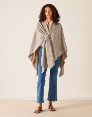 A woman stands indoors in a bright room with light streaming through a large window, smiling softly. She wears her natural curly hair loose and is styled in blue jeans, brown sandals, and a white shirt layered with the MERSEA - MODERN TRAVEL WRAP IN DRIFTWOOD by MERSEA, featuring a securing loop.