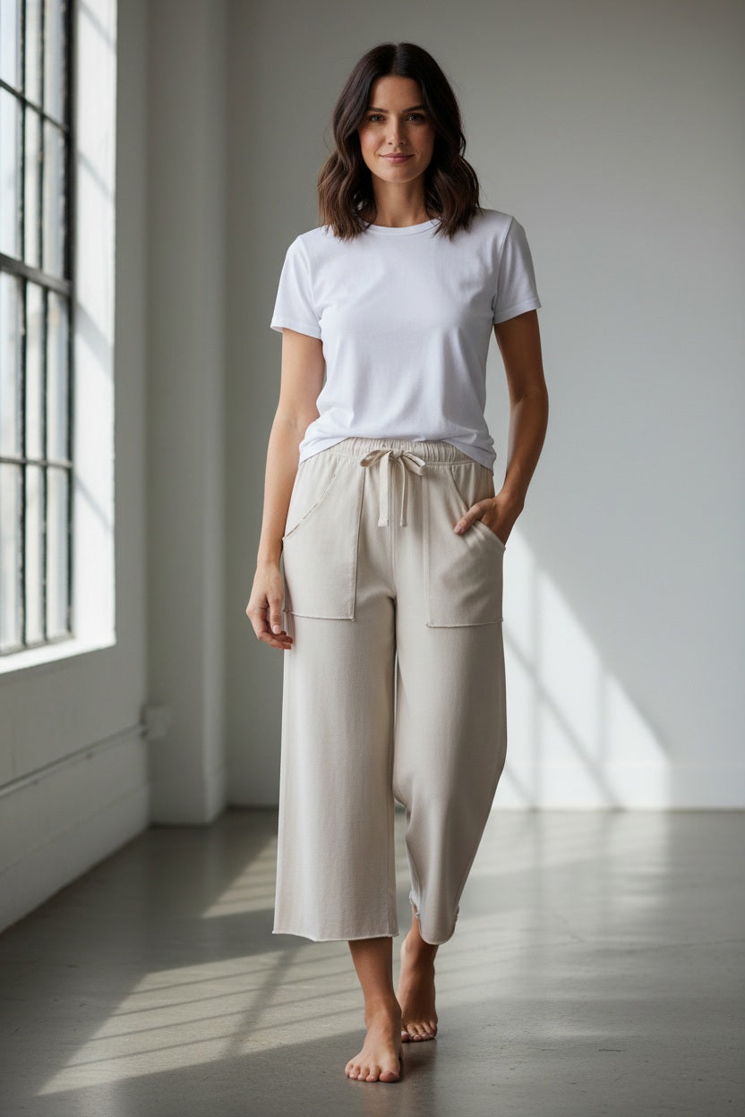 A person wearing the FRANK &amp; EILEEN Penny Patch Pocket Jersey Pant in Vintage White, a striped short-sleeve shirt, and white sandals stands on grass with one hand in their pocket; greenery is in the background.