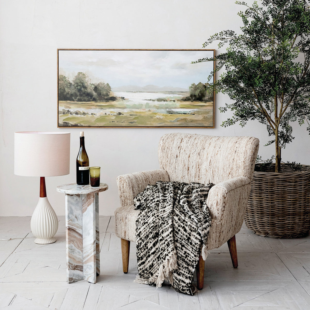 A cozy armchair with a black and white throw blanket sits by a marble side table holding wine. Nearby is the CREATIVE COOP PLEATED STONEWARE TABLE LAMP WITH RUBBERWOOD NECK AND LINEN SHADE, a potted plant, and a landscape painting on the wall.