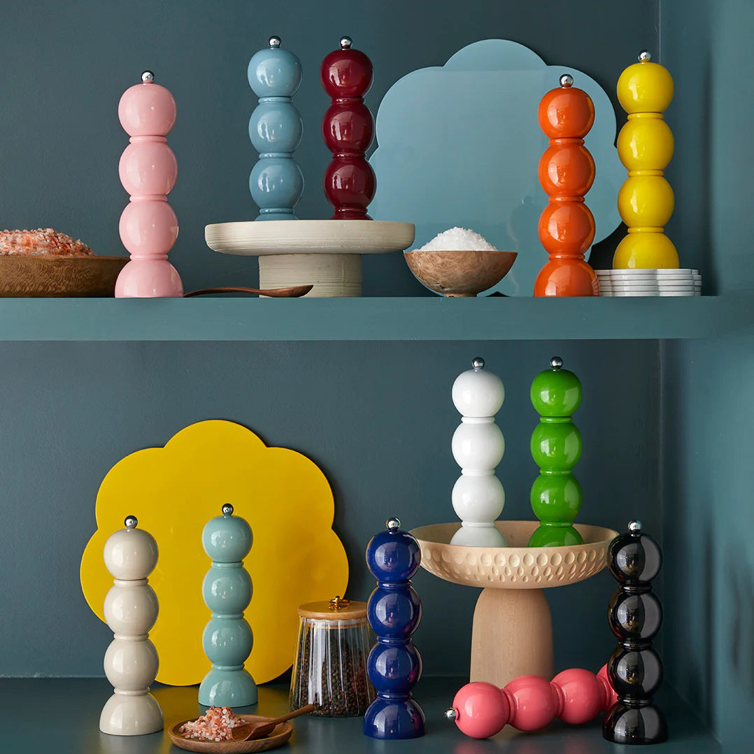ADDISON ROSS&#39;s Bobbin Salt or Pepper Mills, standing at 24cm, are displayed on shelves. These ADDISON ROSS London grinders feature ceramic mechanisms and come in an array of striking color combinations. They are complemented by small bowls filled with salt and seasonings. The muted teal background is adorned with accent shapes that enhance the presentation of the grinders.