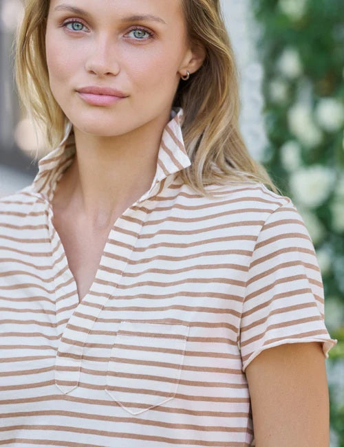 A woman with blonde hair, wearing the FRANK & EILEEN Charlotte Perfect Polo in Heritage Jersey (Vintage White and Camel Stripe) and blue jeans, stands outside near a white building, gazing slightly to the side with one hand gently touching her collar.