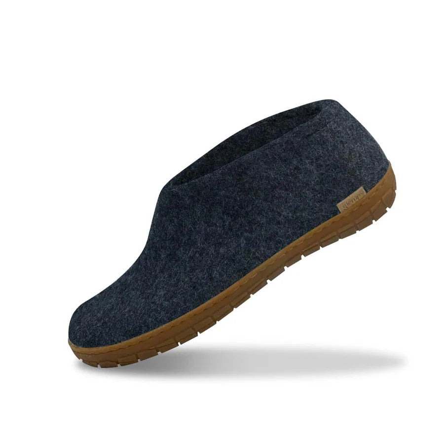A person in a beige coat holds the GLERUPS SHOE WITH NATURAL RUBBER SOLE IN DENIM—cozy, itch-free slippers by GLERUPS, featuring gray natural wool and a brown rubber sole with a branded side label.