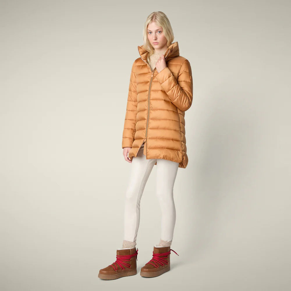 A woman in the SAVE THE DUCK - LYDIA PUFFER COAT by SAVE THE DUCK, paired with a white dress, maroon knee-high boots, and holding a fluffy light pink clutch, stands against a plain beige background.