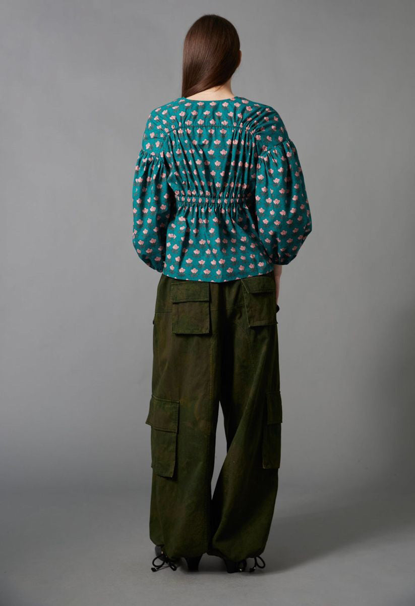 A person with long brown hair, seen from behind, wears the HOPE FOR FLOWERS Shirred Blouse and loose, dark green cargo pants with large pockets, standing against a plain gray background.