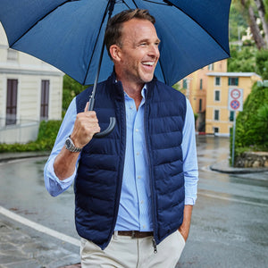 A smiling man in sunglasses and a beige sweater wears the COLLARS AND CO - REGENT REVERSIBLE QUILTED WOOL VEST by COLLARS AND CO, standing before a large decorative fountain in a city square.
