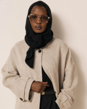 Wearing the HARTFORD - MERA Knit Hood/Scarf in Black—crafted from luxurious wool and cashmere—this person pairs a light long coat with a black turtleneck, wide-leg black pants, and gloves. They stand indoors against a light wall and wooden panel, striking a pose with one hand resting on their face.