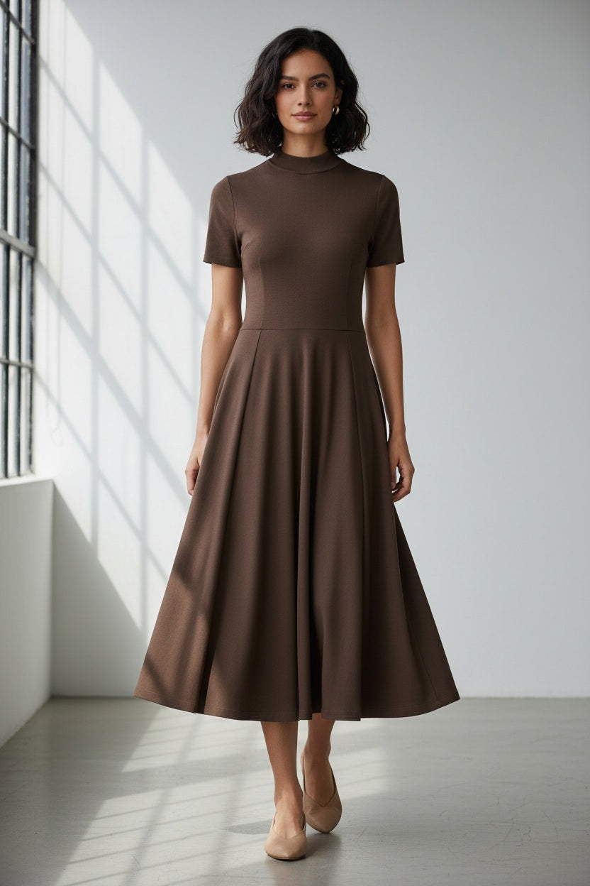 A woman faces away, wearing the LAVI COUTURE - MIDI DEIOPEA DRESS by LAVI in brown. The minimalist design features short sleeves and a fitted waist, styled with a neat bun and heeled sandals against a plain gray background.