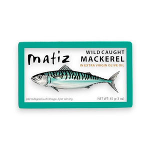 The opened tin of MATIZ - Wild Caught Mackerel in Organic Olive Oil reveals tender fish slices, with an unopened package featuring a mackerel illustration beside it. A fork, crusty bread, and lemon slices are elegantly arranged on a pristine white background. Made in Spain by MATIZ ESPANA with care.