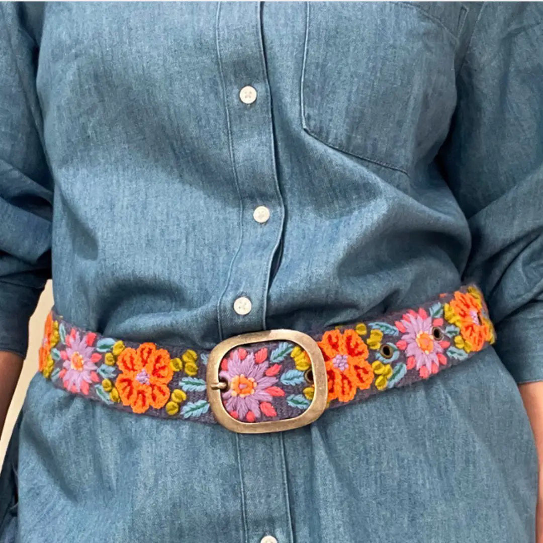 The JENNY KRAUSS - PRAIRIE FLOWERS EMBROIDERED WOOL BELT features colorful floral embroidery in orange, purple, green, and blue on wool, handcrafted in Peru with a silver buckle, shown rolled up on a light surface.