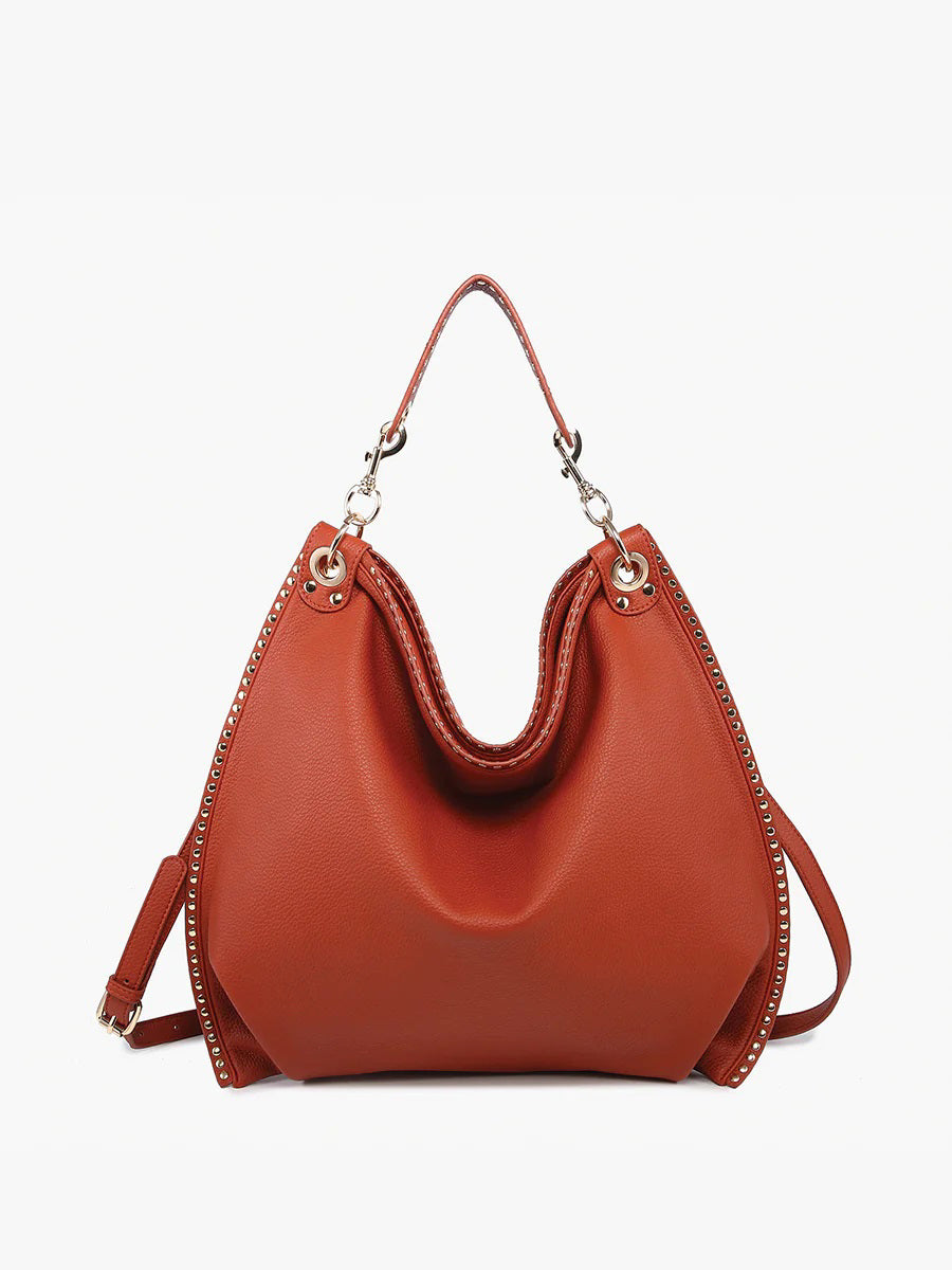 The STUDDED SLOUCHY HOBO by JEN &amp; CO is a brown leather handbag with a slouchy design, featuring a shoulder strap and metal ring accents on the handle attachments. It has a smooth texture and structured shape against a white background.