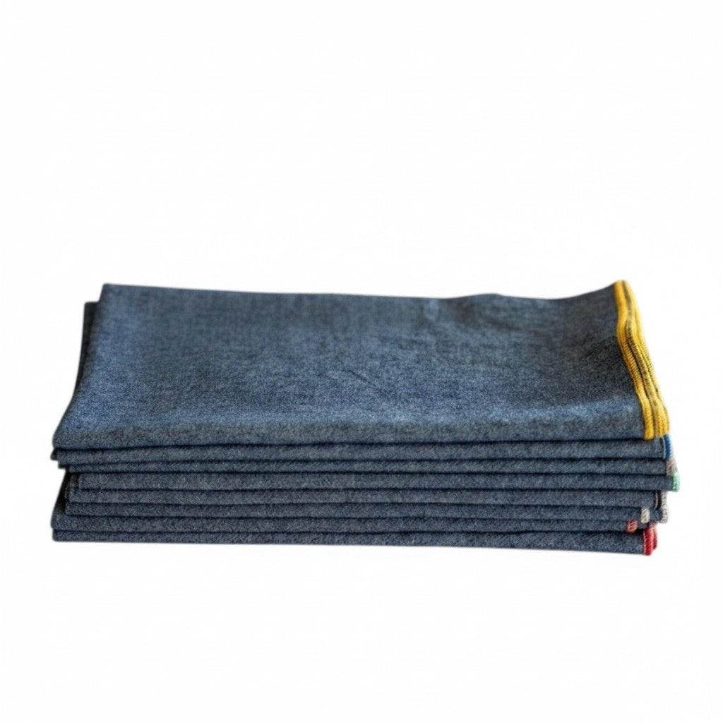 A stack of DOT AND ARMY - CHAMBRAY DINNER NAPKINS rests on a light wooden surface, topped with a sprig of yellow flowers and green leaves.