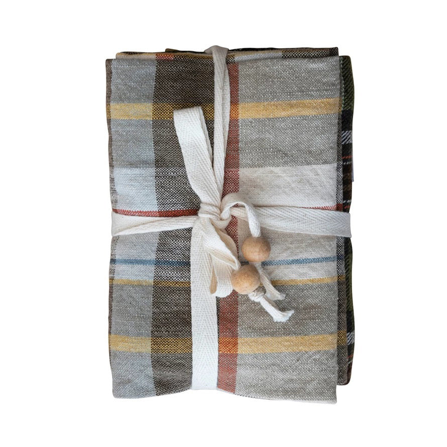 The CREATIVE COOP COTTON PLAID TEA TOWELS - SET OF 3 features brown, beige, yellow, and blue cotton towels folded and tied with a cream fabric ribbon and wooden beads, shown against a white background.