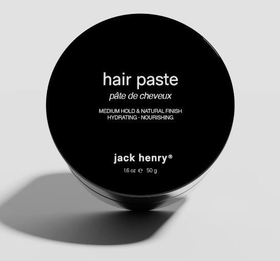 The black JACK HENRY - HAIR PASTE container features "pâte de cheveux" and “jack henry® 16 oz e 50 g” on the lid, offering medium hold, hydrating nourishment, and natural shine for healthy hair growth against a light background.