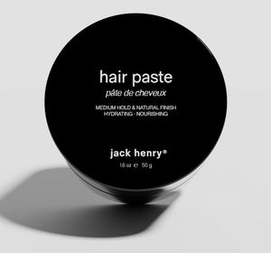The black JACK HENRY - HAIR PASTE container features "pâte de cheveux" and “jack henry® 16 oz e 50 g” on the lid, offering medium hold, hydrating nourishment, and natural shine for healthy hair growth against a light background.