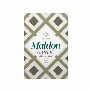A box of GOURMET FOOD SOLUTIONS' MALDON - GARLIC SEA SALT is placed on a bed of coarse salt with herbs and garlic flakes scattered around. The box, in a green and white geometric design, reads "MALDON - GARLIC SEA SALT" and "with white & roasted garlic," established 1882—proudly showcasing its English sea salt heritage.