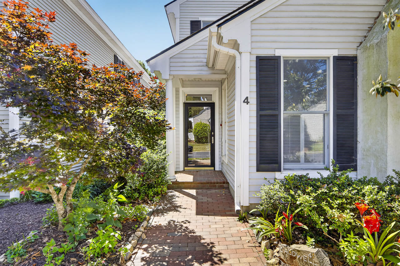 A brick walkway leads to the glass front door of a white house with black shutters. Lush green plants and a small tree line the path under a clear, sunny sky.