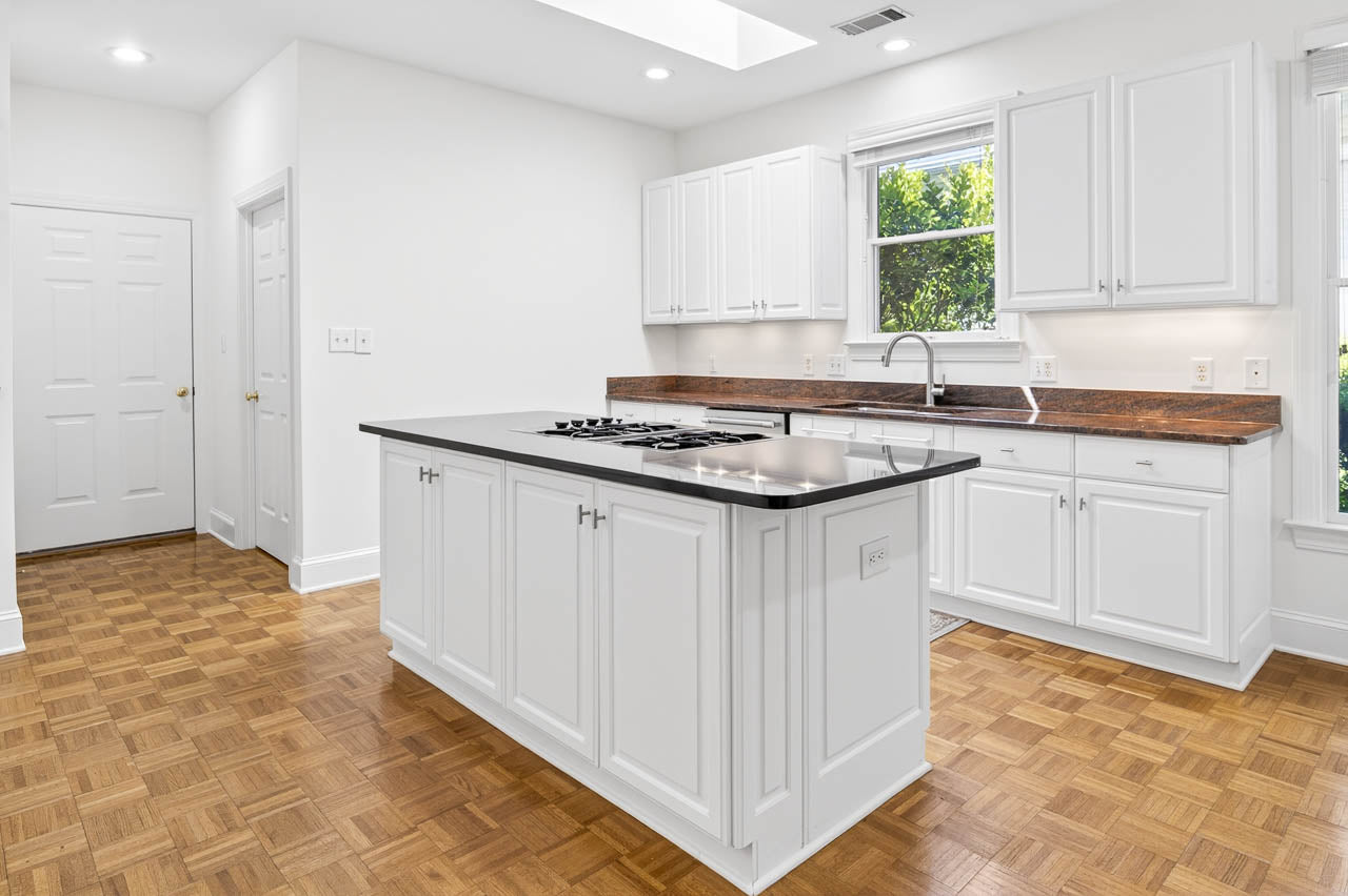 A bright, modern kitchen with white cabinets, a black countertop island with a built-in stovetop, wood parquet flooring, large windows, and ample natural light.