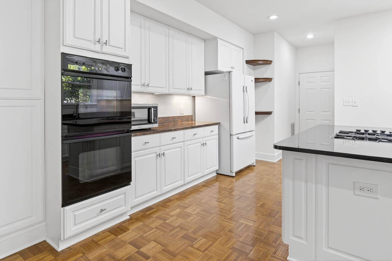 Modern kitchen with white cabinets, double wall oven, countertop microwave, white refrigerator, gas cooktop on island, wood parquet flooring, and open shelves in the corner. Room is well-lit with recessed lighting.
